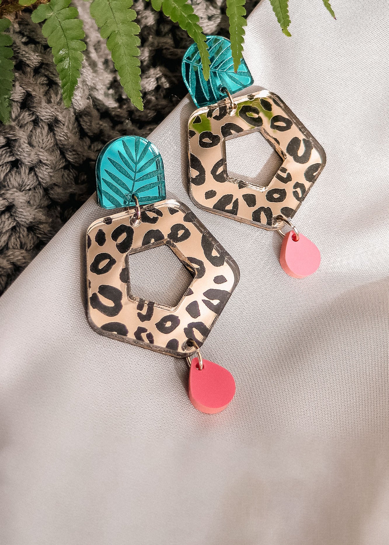 A pair of Leopard Earrings featuring leopard print hexagons, turquoise and black leaf-patterned tops, and dangling pink teardrops—crafted from premium materials, displayed on white fabric with a green fern leaf nearby.