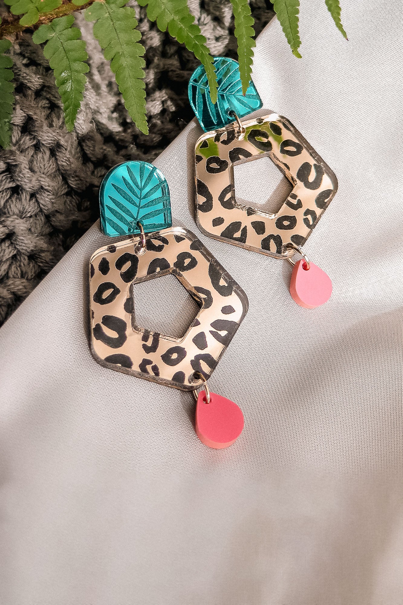 A pair of Leopard Earrings featuring leopard print hexagons, turquoise and black leaf-patterned tops, and dangling pink teardrops—crafted from premium materials, displayed on white fabric with a green fern leaf nearby.