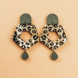 A pair of Leopard Earrings with olive green tops and teardrop-shaped dangles, featuring hexagonal centers in timeless patterns, crafted from premium materials and displayed on a peach background.