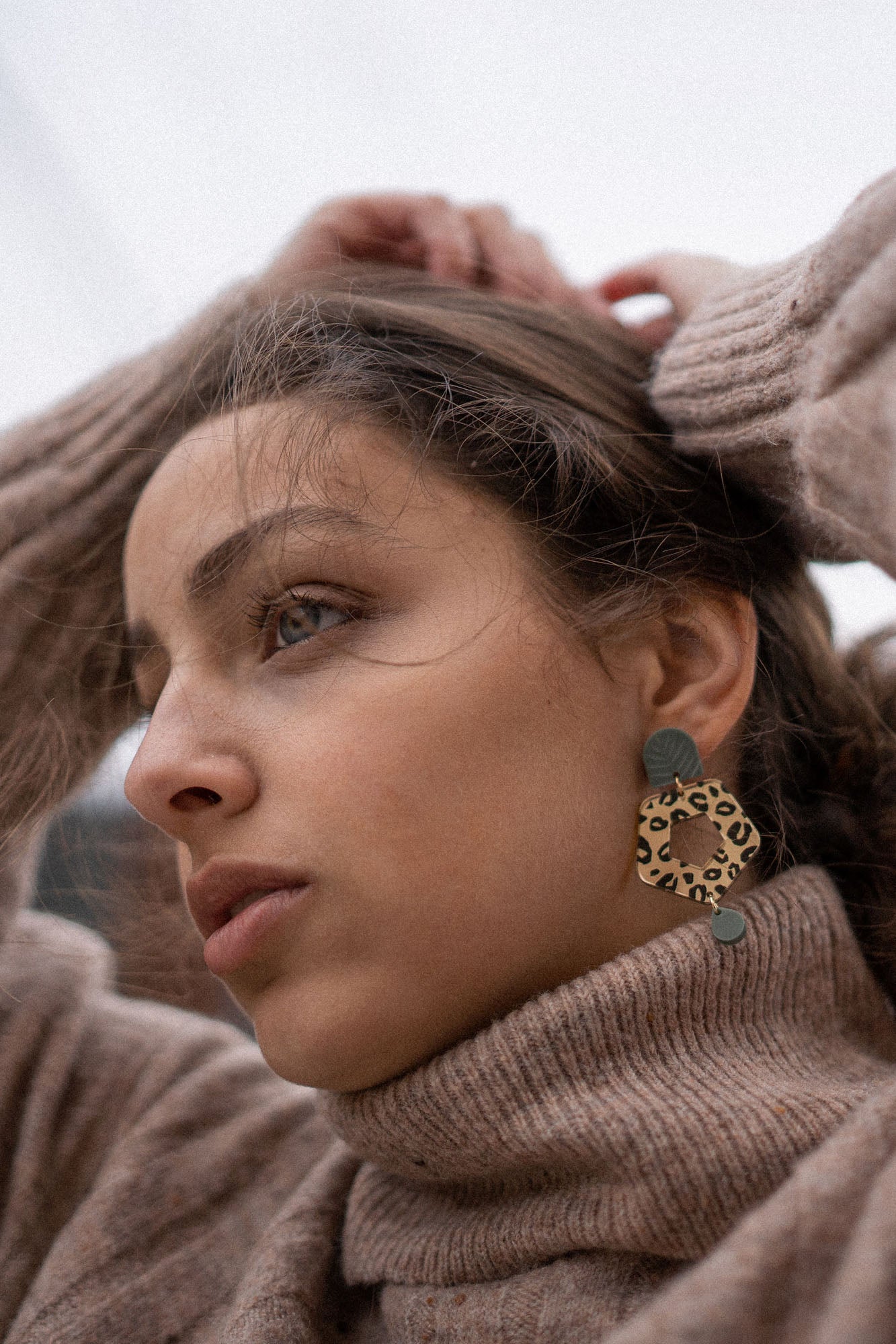 A woman in a brown turtleneck sweater gazes thoughtfully to the side, hands raised behind her head. She wears Leopard Earrings made from premium materials, their timeless patterns adding bold elegance to her look.