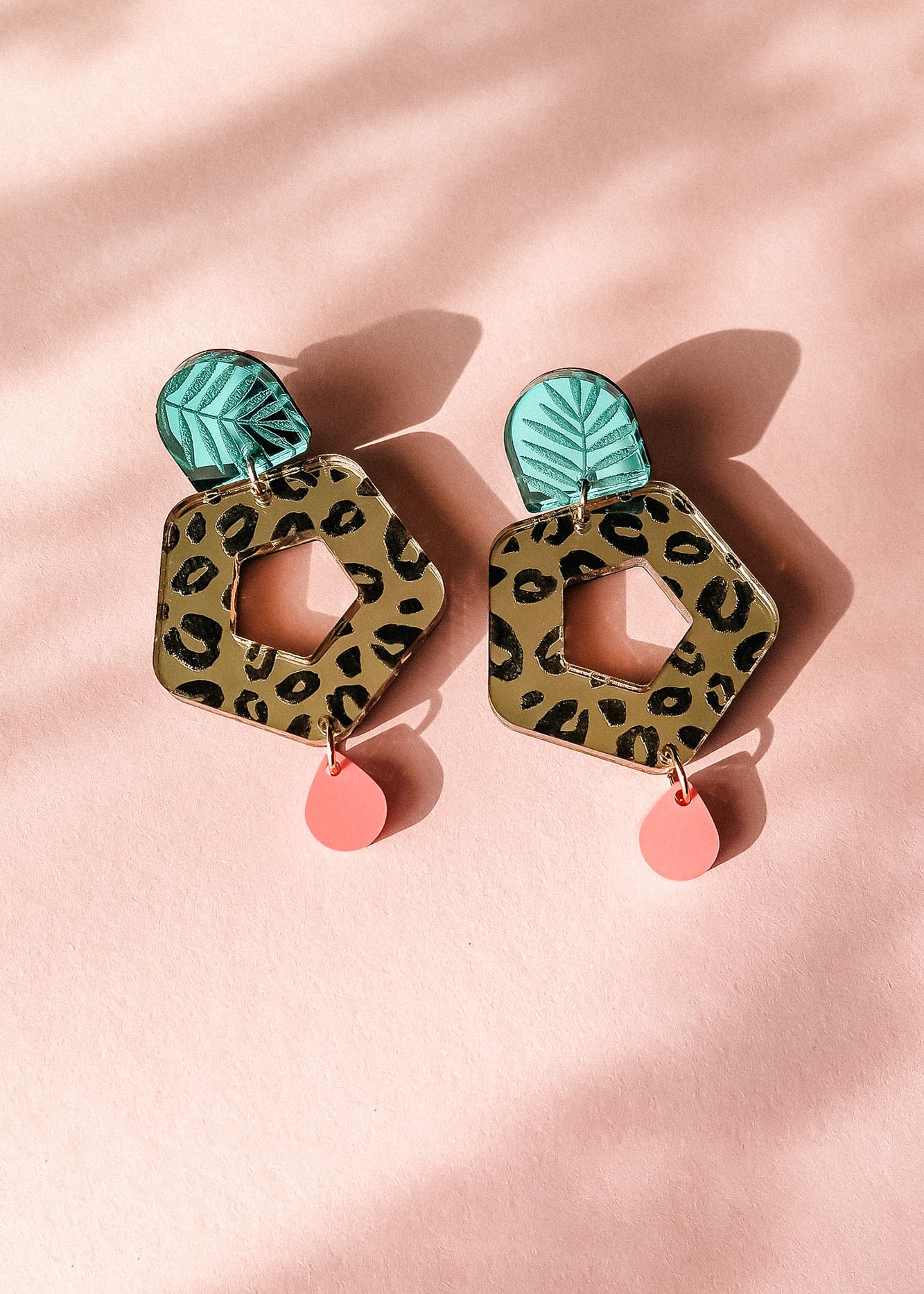 A pair of Leopard Earrings featuring green leaf-shaped tops, hexagonal leopard-print centers with cutouts, and small pink teardrop dangles—displayed on a light pink background for standout style with timeless patterns.