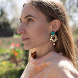 A woman with light brown hair gazes to the left, wearing a peach top and striking Leopard Earrings crafted from premium materials. The background is outdoors with blurred greenery and flowers.