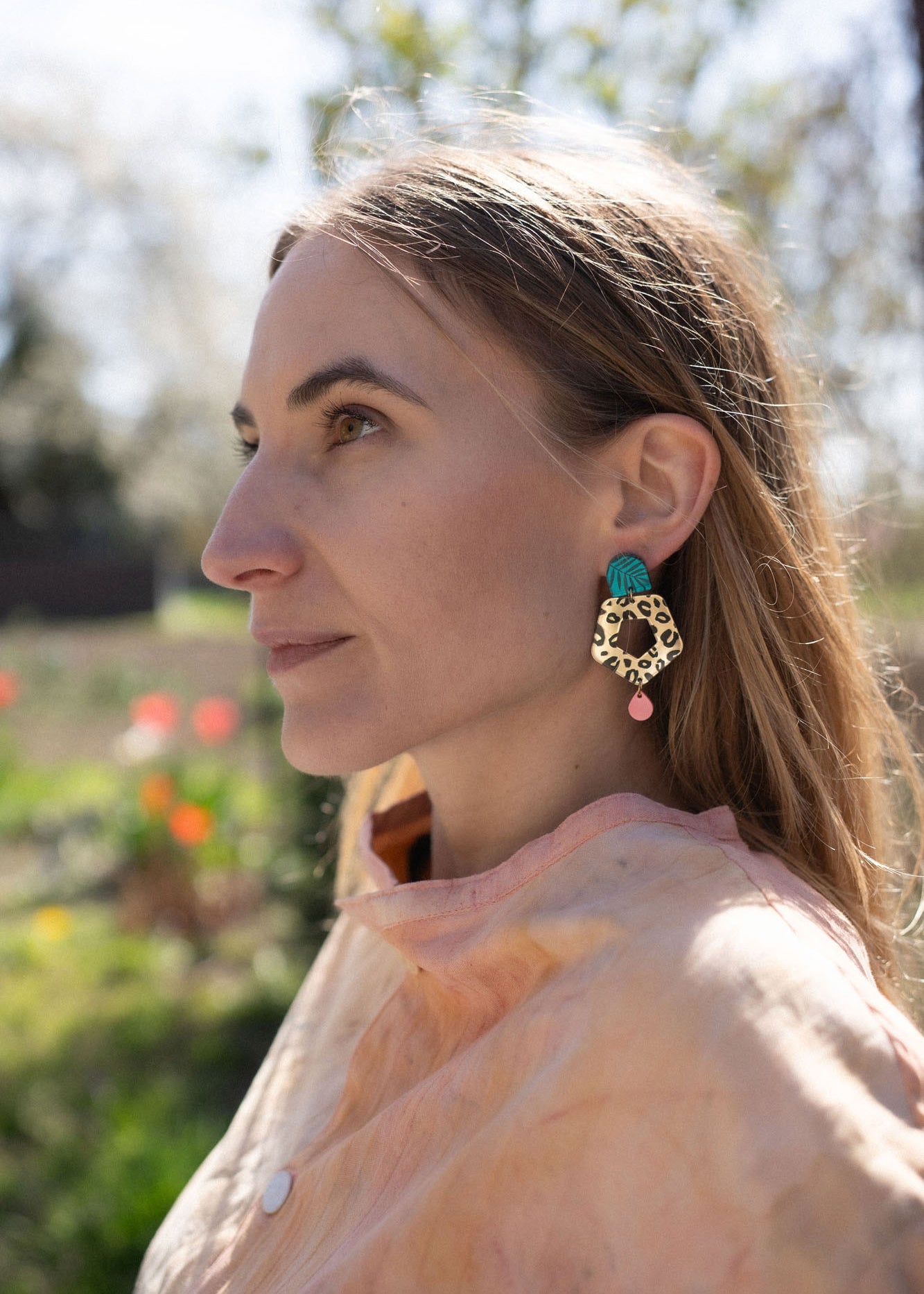 A woman with light brown hair gazes to the left, wearing a peach top and striking Leopard Earrings crafted from premium materials. The background is outdoors with blurred greenery and flowers.