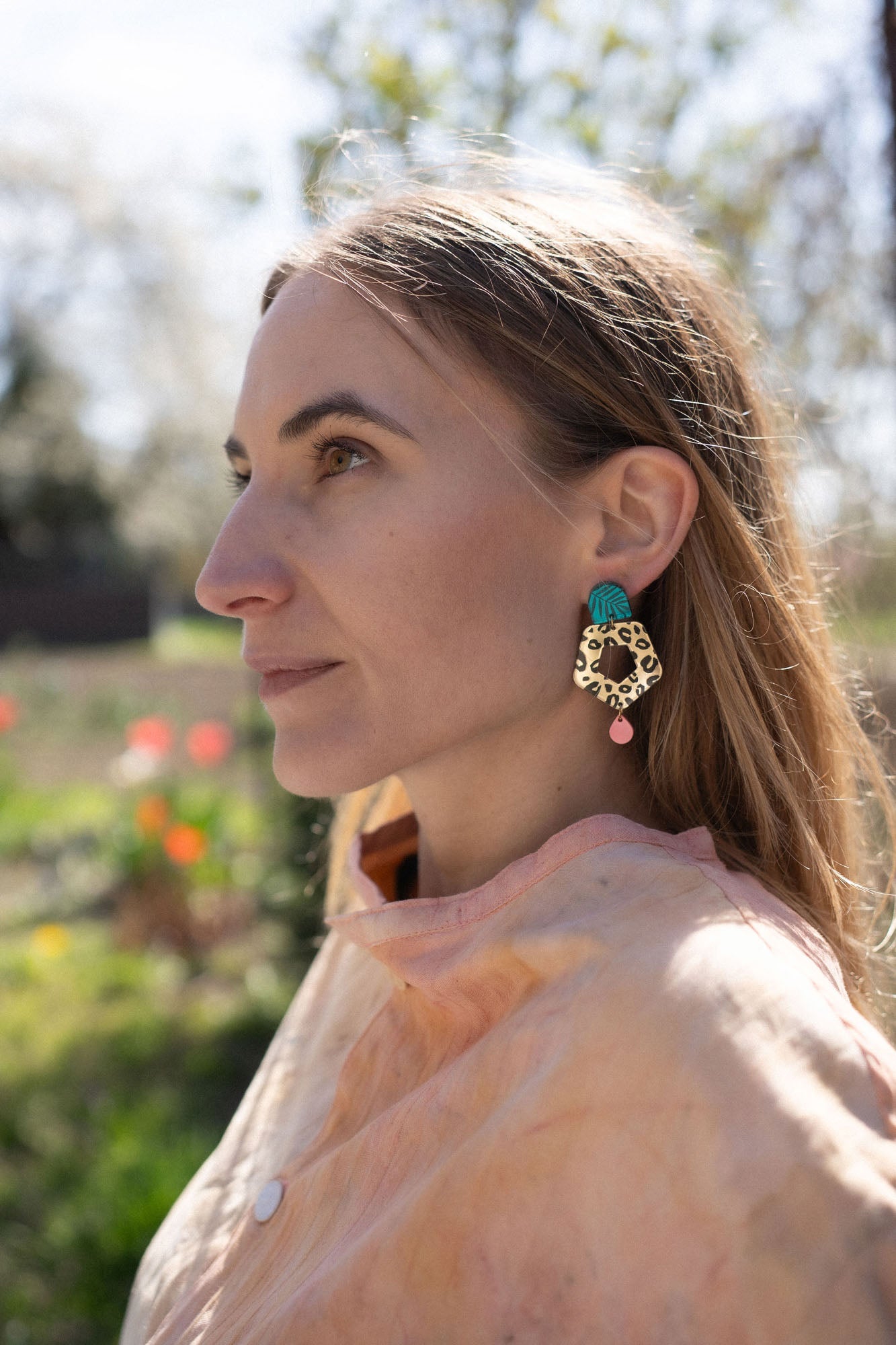 A woman with light brown hair gazes to the left, wearing a peach top and striking Leopard Earrings crafted from premium materials. The background is outdoors with blurred greenery and flowers.