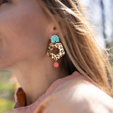 A close-up of a woman’s face in profile, wearing Leopard Earrings featuring timeless patterns—a large, colorful leopard print with a blue geometric shape and a small red drop—paired with a light peach top outdoors in sunlight.