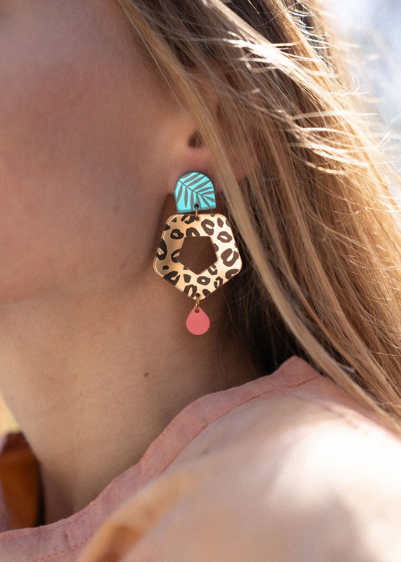 A close-up of a woman’s face in profile, wearing Leopard Earrings featuring timeless patterns—a large, colorful leopard print with a blue geometric shape and a small red drop—paired with a light peach top outdoors in sunlight.