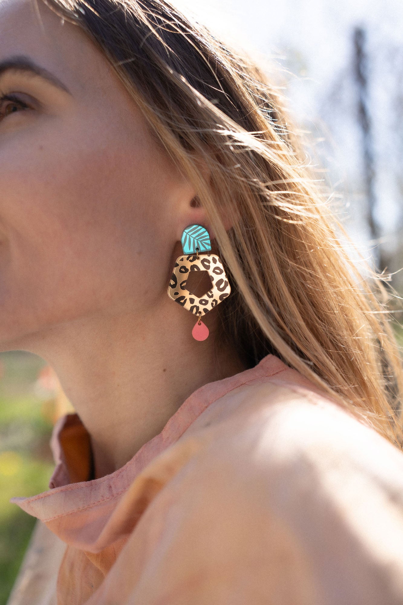 A close-up of a woman’s face in profile, wearing Leopard Earrings featuring timeless patterns—a large, colorful leopard print with a blue geometric shape and a small red drop—paired with a light peach top outdoors in sunlight.