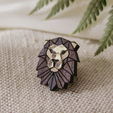 A handcrafted geometric lion pin with metallic gold and dark purple facets rests on textured beige fabric, complemented by green fern leaves in the background.