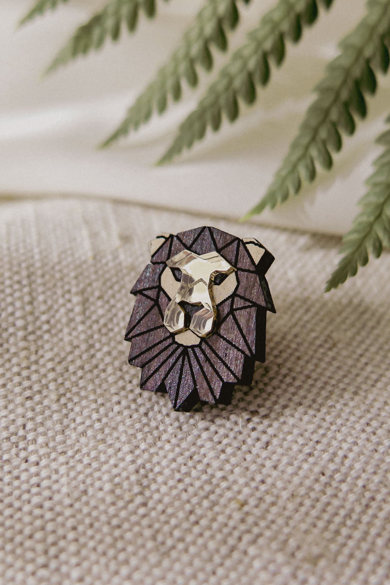 A handcrafted geometric lion pin with metallic gold and dark purple facets rests on textured beige fabric, complemented by green fern leaves in the background.