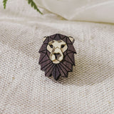 A handcrafted geometric Lion Pin brooch with a dark mane and gold face rests on textured cream fabric, with green fern leaves partially visible above.