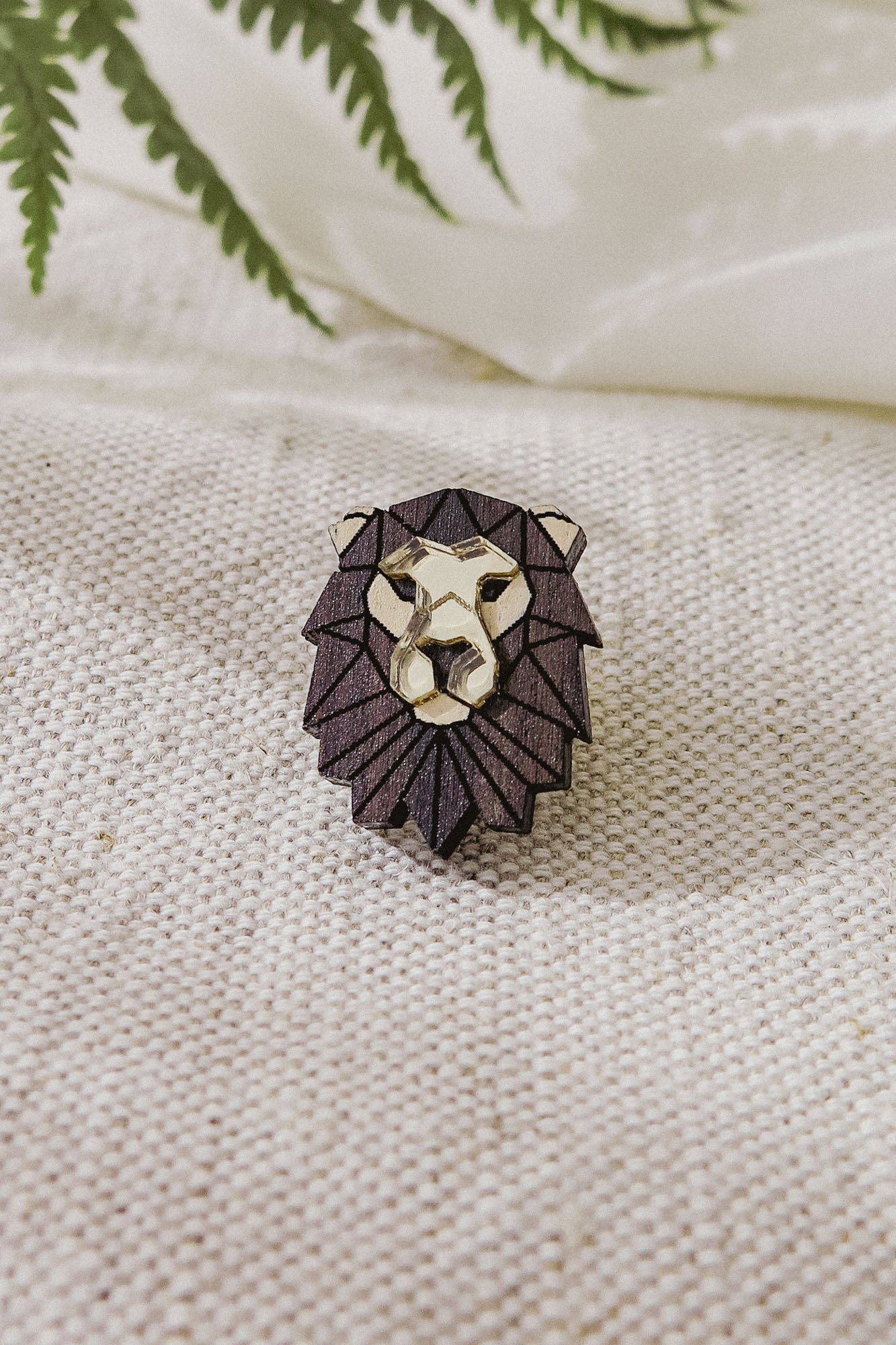 A handcrafted geometric Lion Pin brooch with a dark mane and gold face rests on textured cream fabric, with green fern leaves partially visible above.
