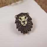 A handcrafted geometric Lion Pin made of dark wood with gold highlights, placed on a white surface with dried grasses in the background.