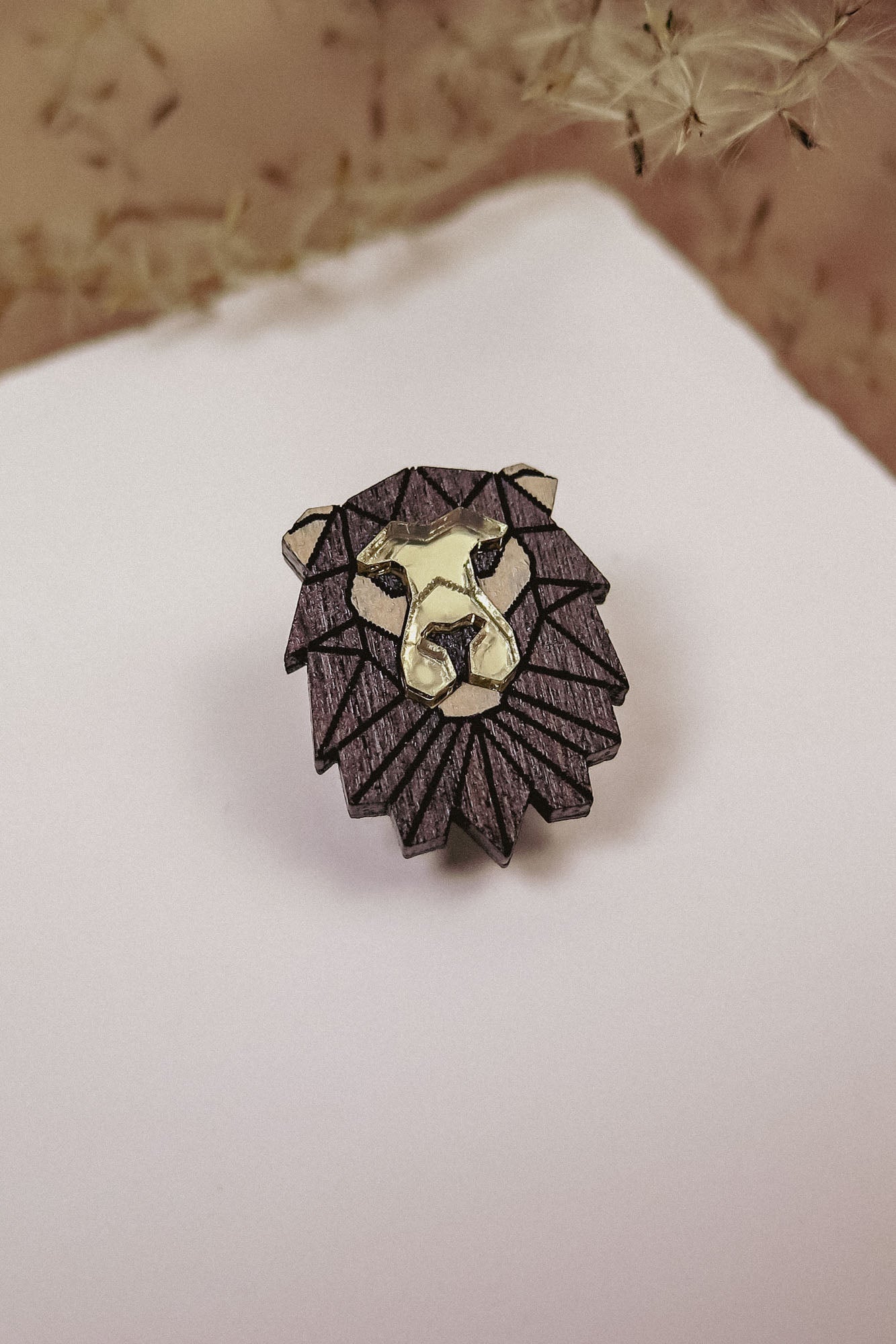 A handcrafted geometric Lion Pin made of dark wood with gold highlights, placed on a white surface with dried grasses in the background.