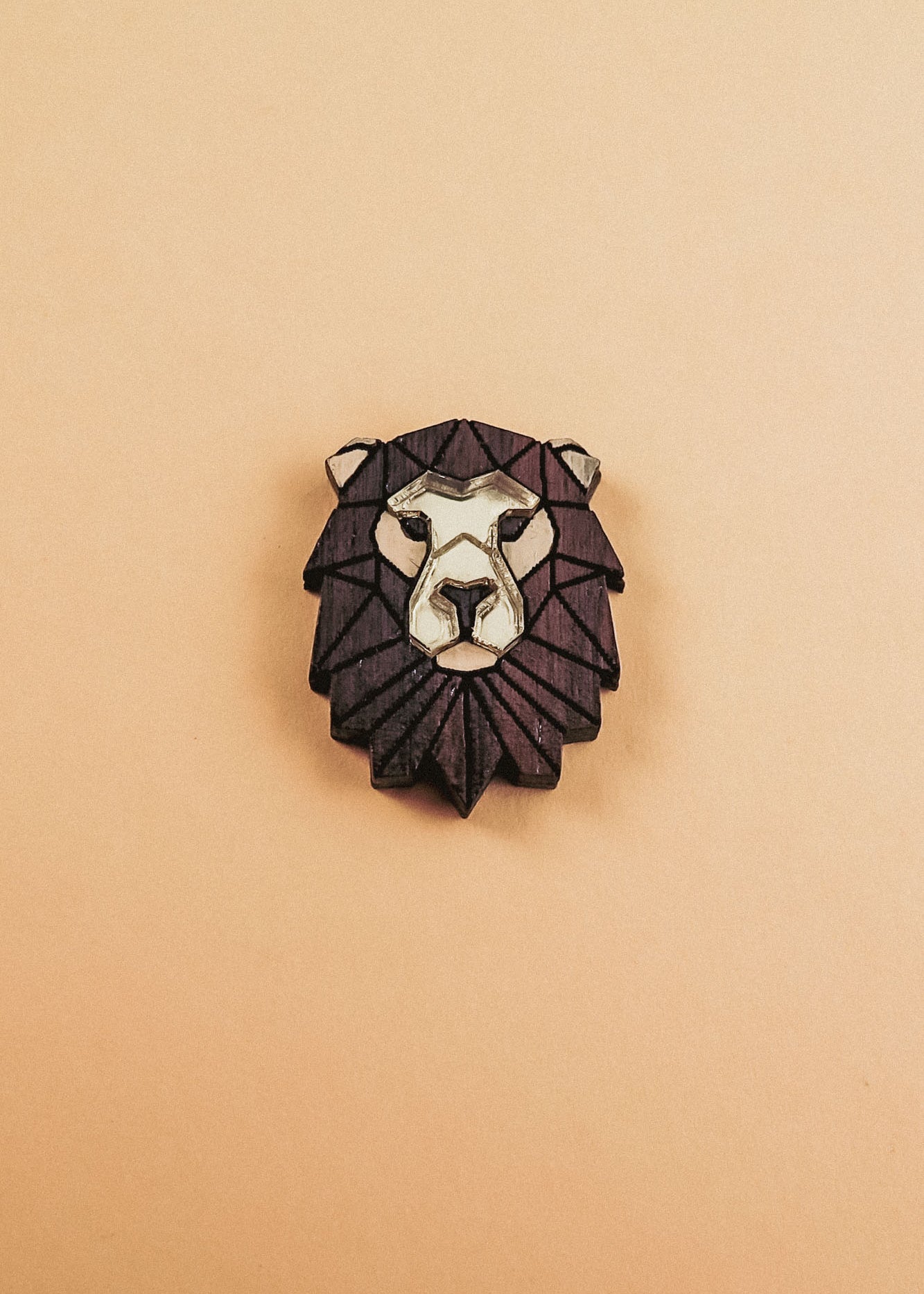 A handcrafted geometric Lion Pin brooch with a dark brown mane and light beige face, displayed on a plain light tan background.