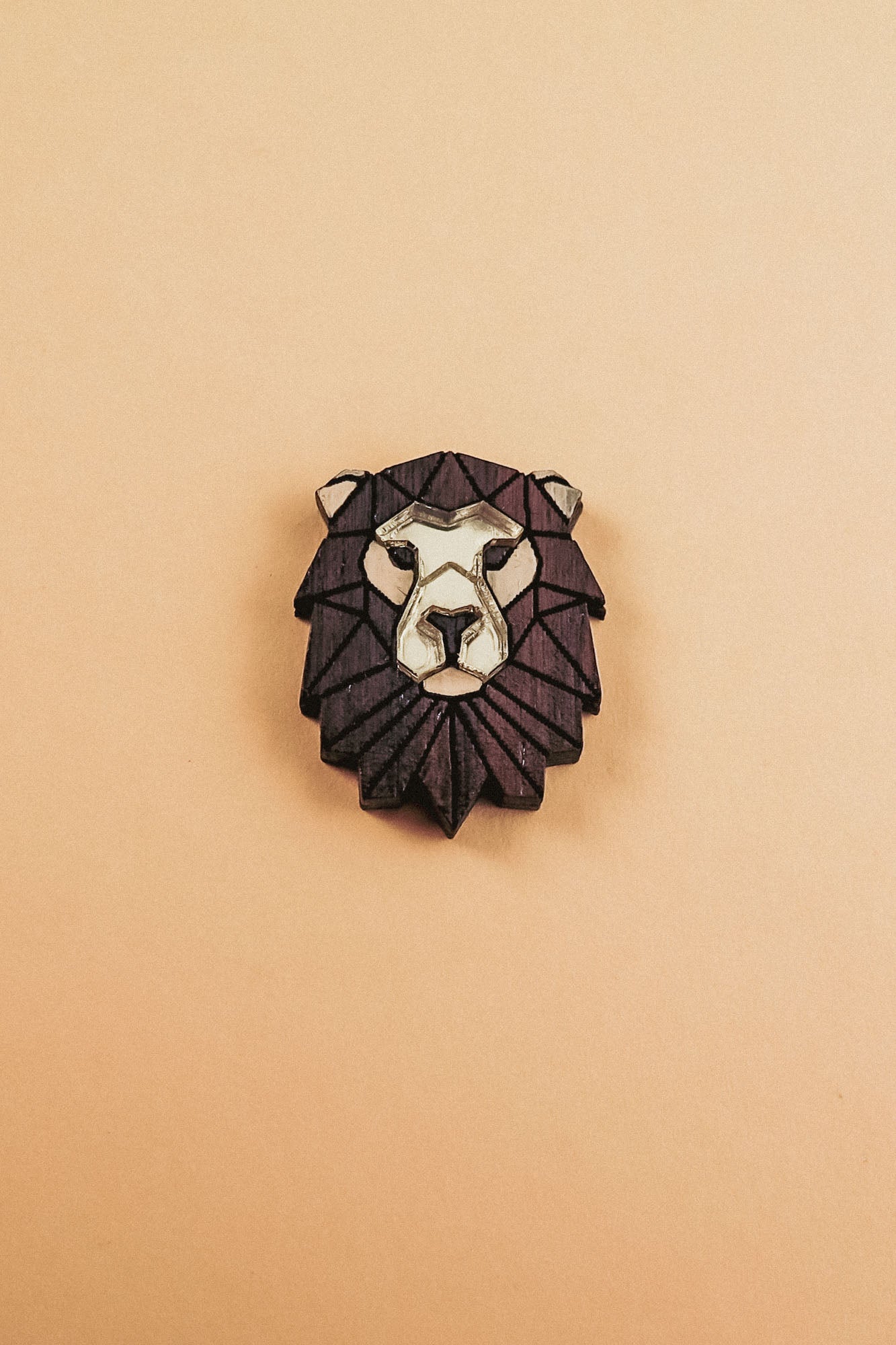 A handcrafted geometric Lion Pin brooch with a dark brown mane and light beige face, displayed on a plain light tan background.