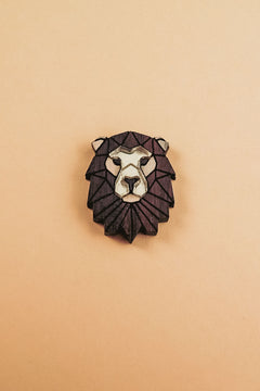 A handcrafted geometric Lion Pin brooch with a dark brown mane and light beige face, displayed on a plain light tan background.