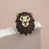 A handcrafted Lion Pin brooch with gold and dark brown accents rests on the edge of a white textured surface against a soft pink background, serving as a striking geometric accessory.