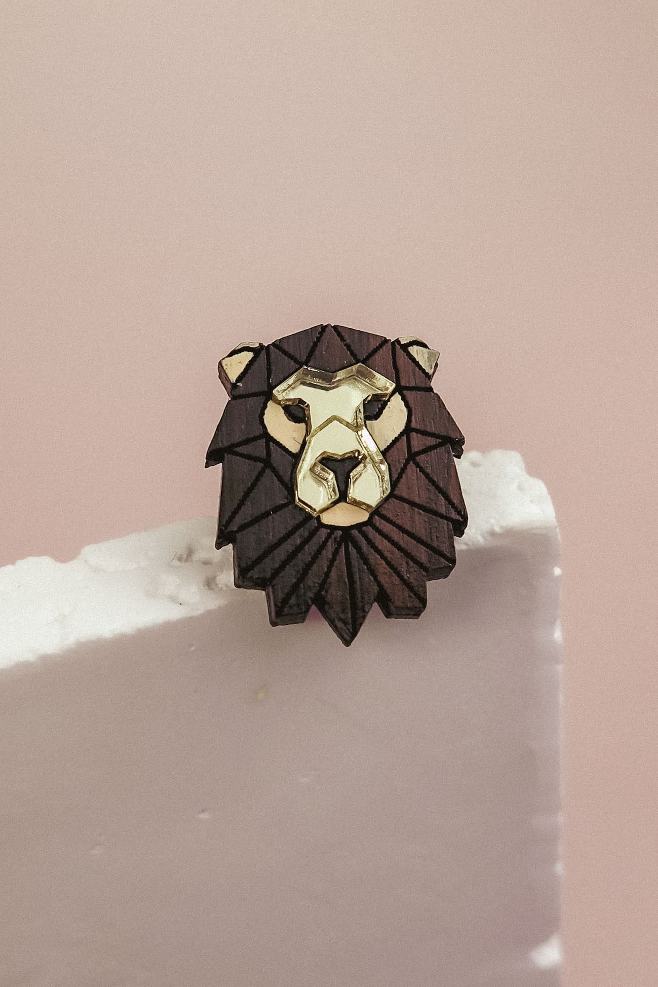 A handcrafted Lion Pin brooch with gold and dark brown accents rests on the edge of a white textured surface against a soft pink background, serving as a striking geometric accessory.