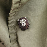 A close-up of an olive green jacket with a handcrafted geometric accessory—a Lion Pin—attached near a button.