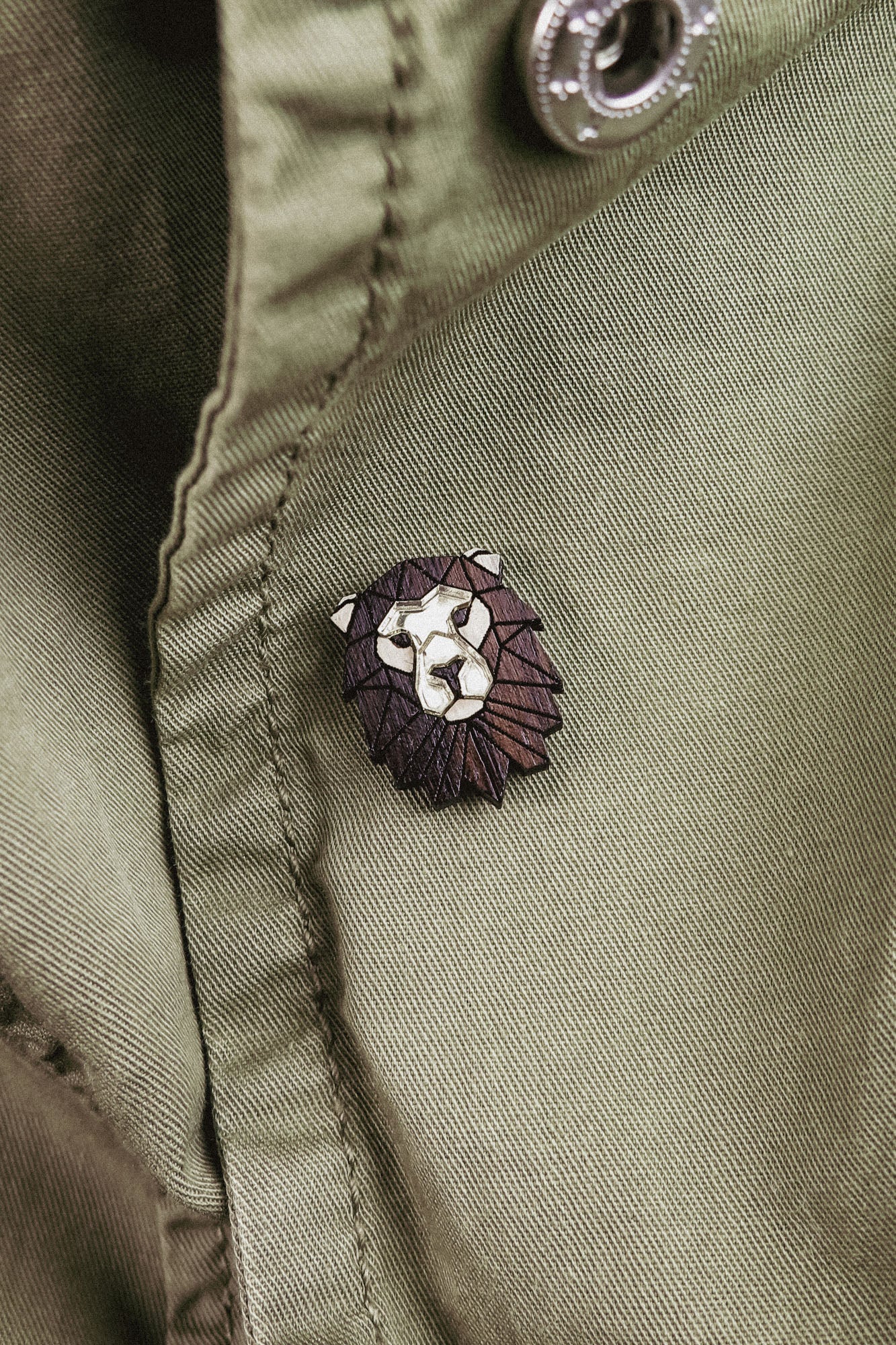 A close-up of an olive green jacket with a handcrafted geometric accessory—a Lion Pin—attached near a button.