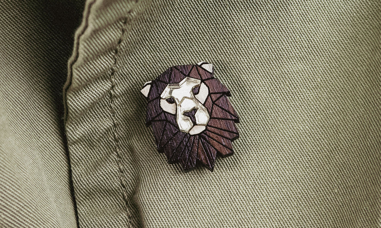 A close-up of an olive green jacket with a handcrafted geometric accessory—a Lion Pin—attached near a button.