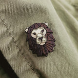 A handcrafted metallic lion pin with a dark, geometric mane detail is attached to a light olive green fabric, possibly a jacket.