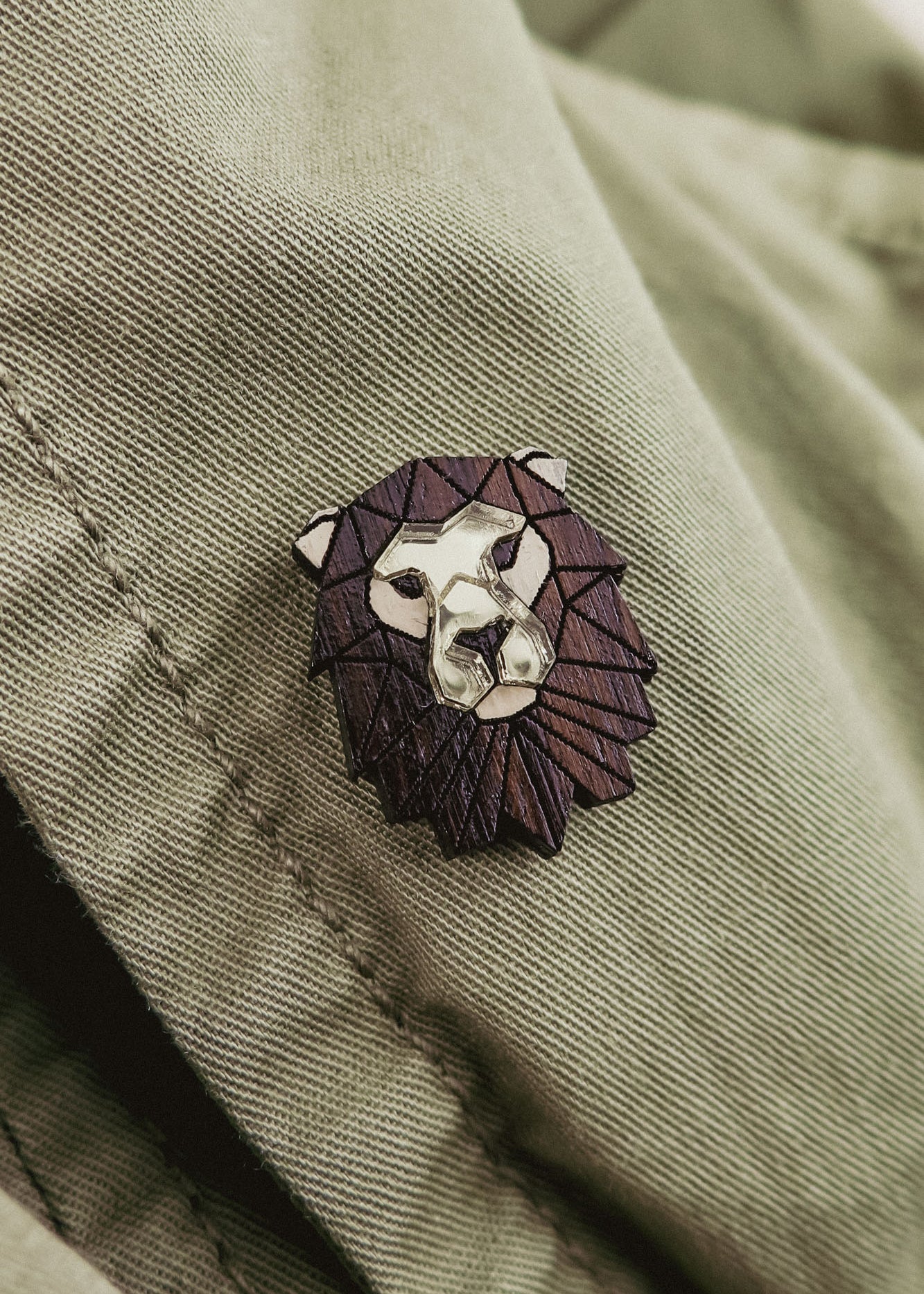 A handcrafted metallic lion pin with a dark, geometric mane detail is attached to a light olive green fabric, possibly a jacket.