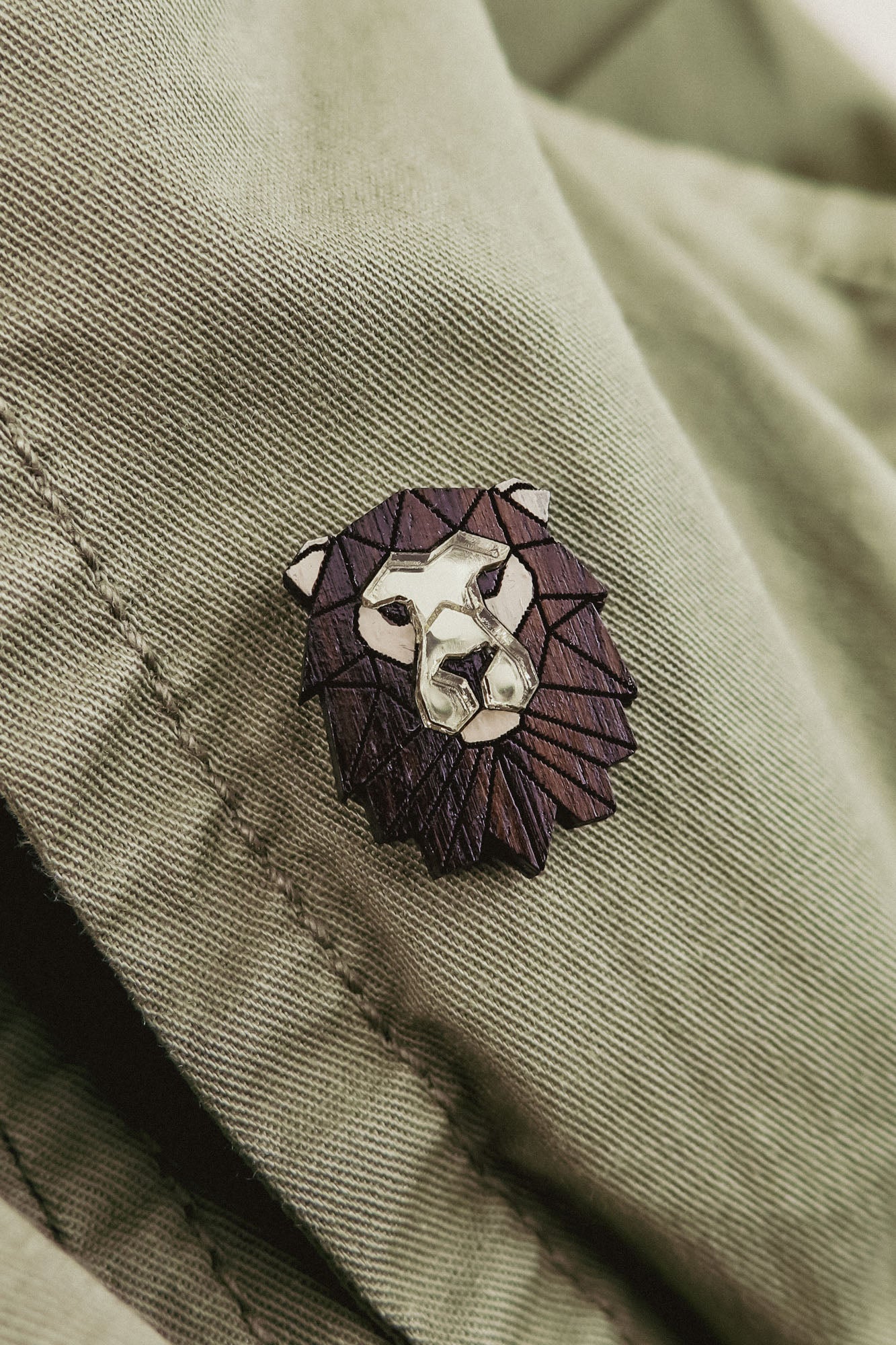 A handcrafted metallic lion pin with a dark, geometric mane detail is attached to a light olive green fabric, possibly a jacket.