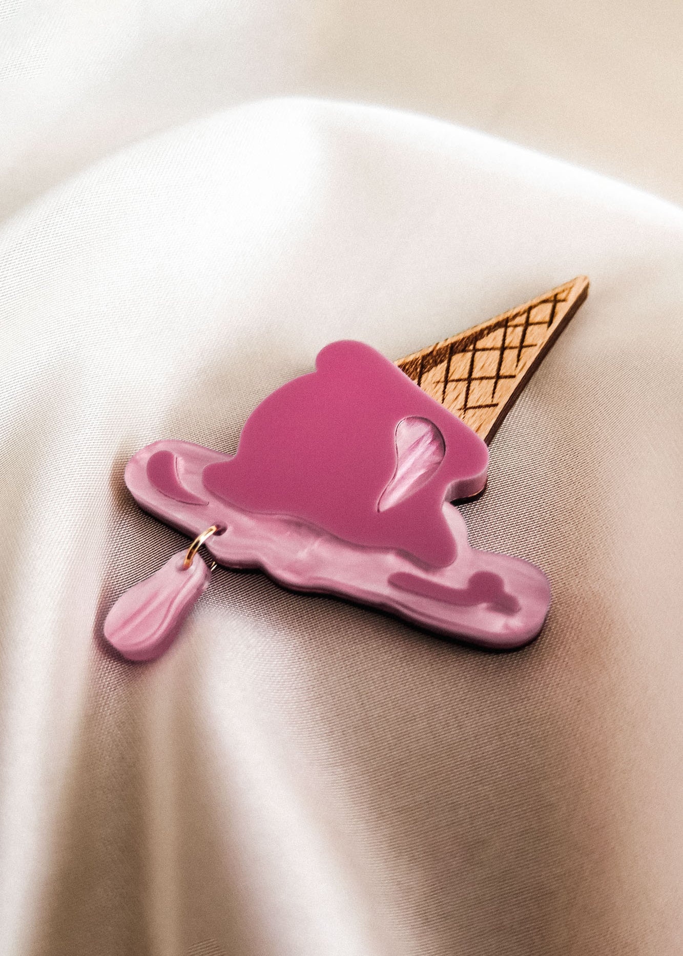 A handcrafted Melted Ice Cream Brooch, shaped like a melting pink scoop on a waffle cone, rests on a smooth, cream-colored fabric background—perfect for adding charm to your summer wardrobe.