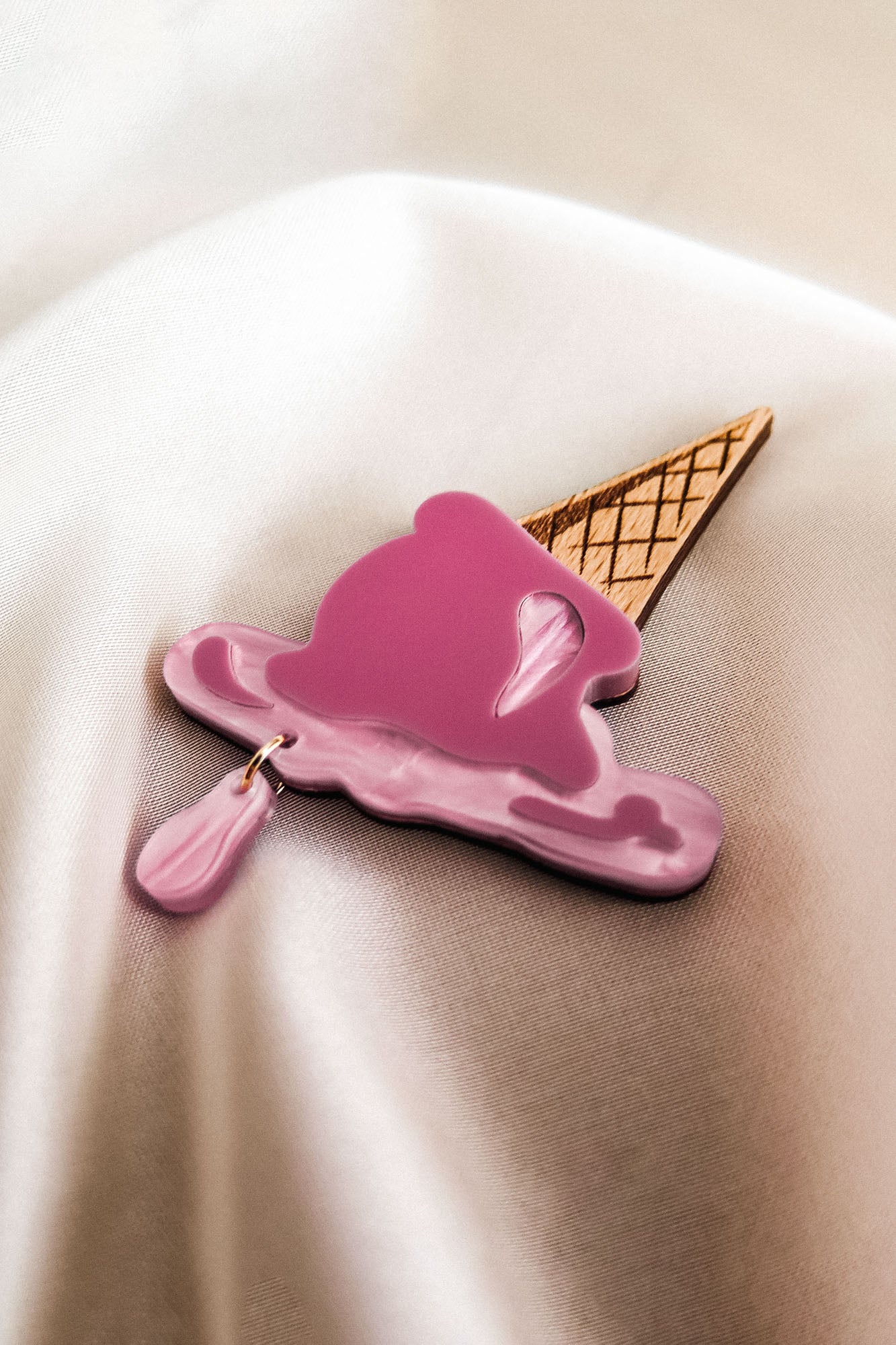 A handcrafted Melted Ice Cream Brooch, shaped like a melting pink scoop on a waffle cone, rests on a smooth, cream-colored fabric background—perfect for adding charm to your summer wardrobe.