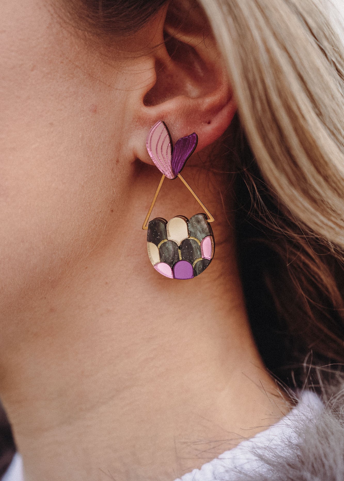 Close-up of a woman's ear wearing playful Mermaid Tail Earrings with a pink and purple heart-shaped stud and a semi-circular dangle in green, pink, and purple—a perfect accessory for mermaid lovers. The woman has blonde hair and wears a white top.