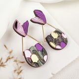 A pair of colorful drop earrings with abstract petal shapes in purple, pink, yellow, and green, displayed on a white textured surface—perfect for mermaid lovers seeking whimsical elegance in their accessory collection.
