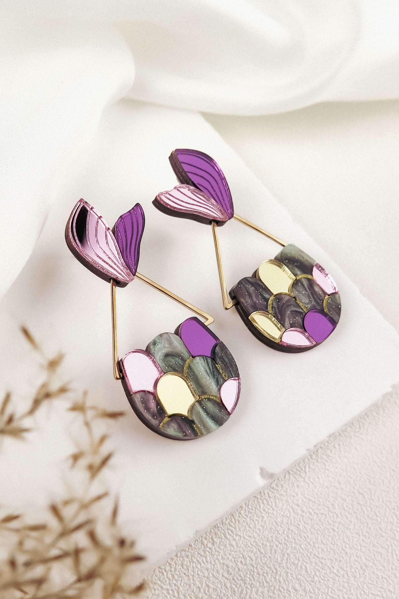 A pair of colorful drop earrings with abstract petal shapes in purple, pink, yellow, and green, displayed on a white textured surface—perfect for mermaid lovers seeking whimsical elegance in their accessory collection.