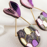 Colorful Mermaid Tail Earrings with a fan-shaped purple top and a semi-circular base featuring metallic and pastel scales in gold, pink, and gray, perfect for mermaid lovers seeking whimsical elegance. Displayed on a soft, textured white surface.