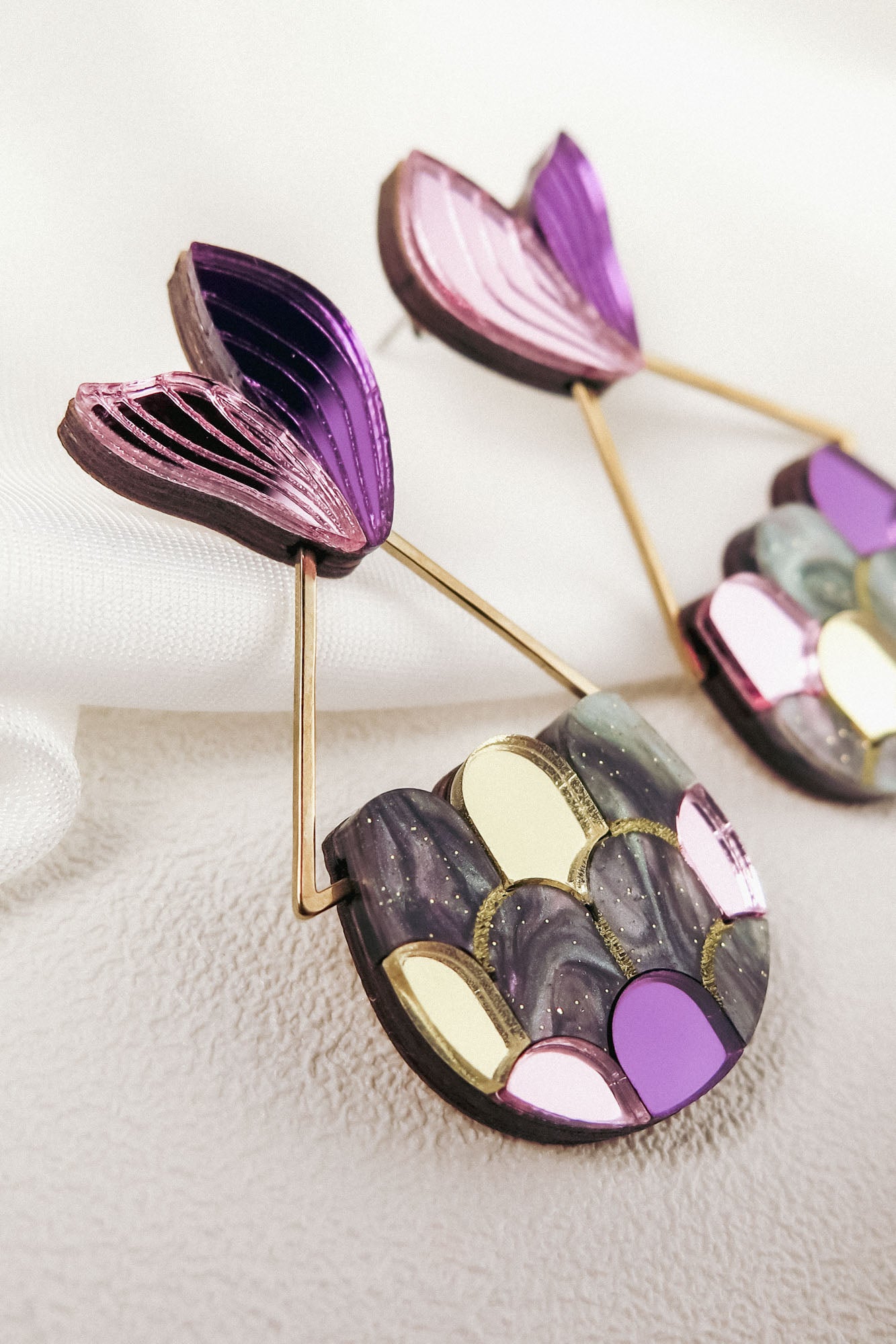 Colorful Mermaid Tail Earrings with a fan-shaped purple top and a semi-circular base featuring metallic and pastel scales in gold, pink, and gray, perfect for mermaid lovers seeking whimsical elegance. Displayed on a soft, textured white surface.