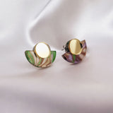 A pair of minimalist geometric stud earrings featuring round gold studs and semicircular marbled green and purple acrylic accents, displayed on soft, white fabric.