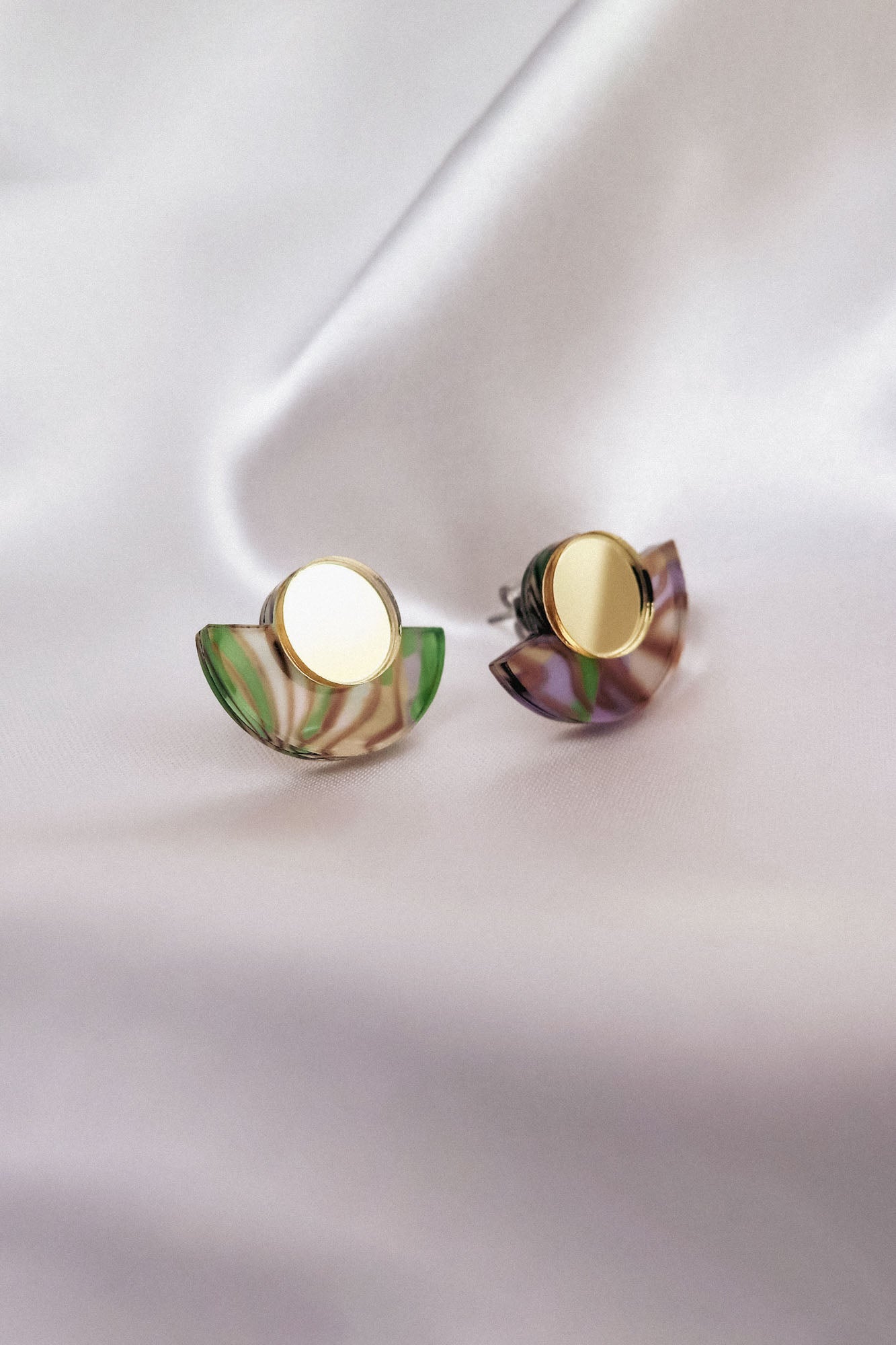 A pair of minimalist geometric stud earrings featuring round gold studs and semicircular marbled green and purple acrylic accents, displayed on soft, white fabric.