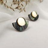 A pair of minimalist geometric stud earrings featuring a gold mirrored circle atop a dark, semi-circular marbled base, displayed on a white surface with dried plant accents in the background.