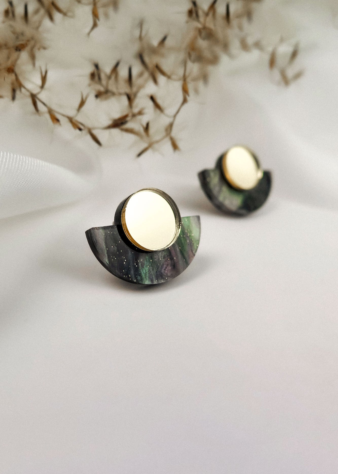 A pair of minimalist geometric stud earrings featuring a gold mirrored circle atop a dark, semi-circular marbled base, displayed on a white surface with dried plant accents in the background.