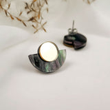A pair of minimalist geometric stud earrings featuring a circular gold piece atop a semi-circular, marbled purple and green base, displayed on a white surface with dried foliage in the background.