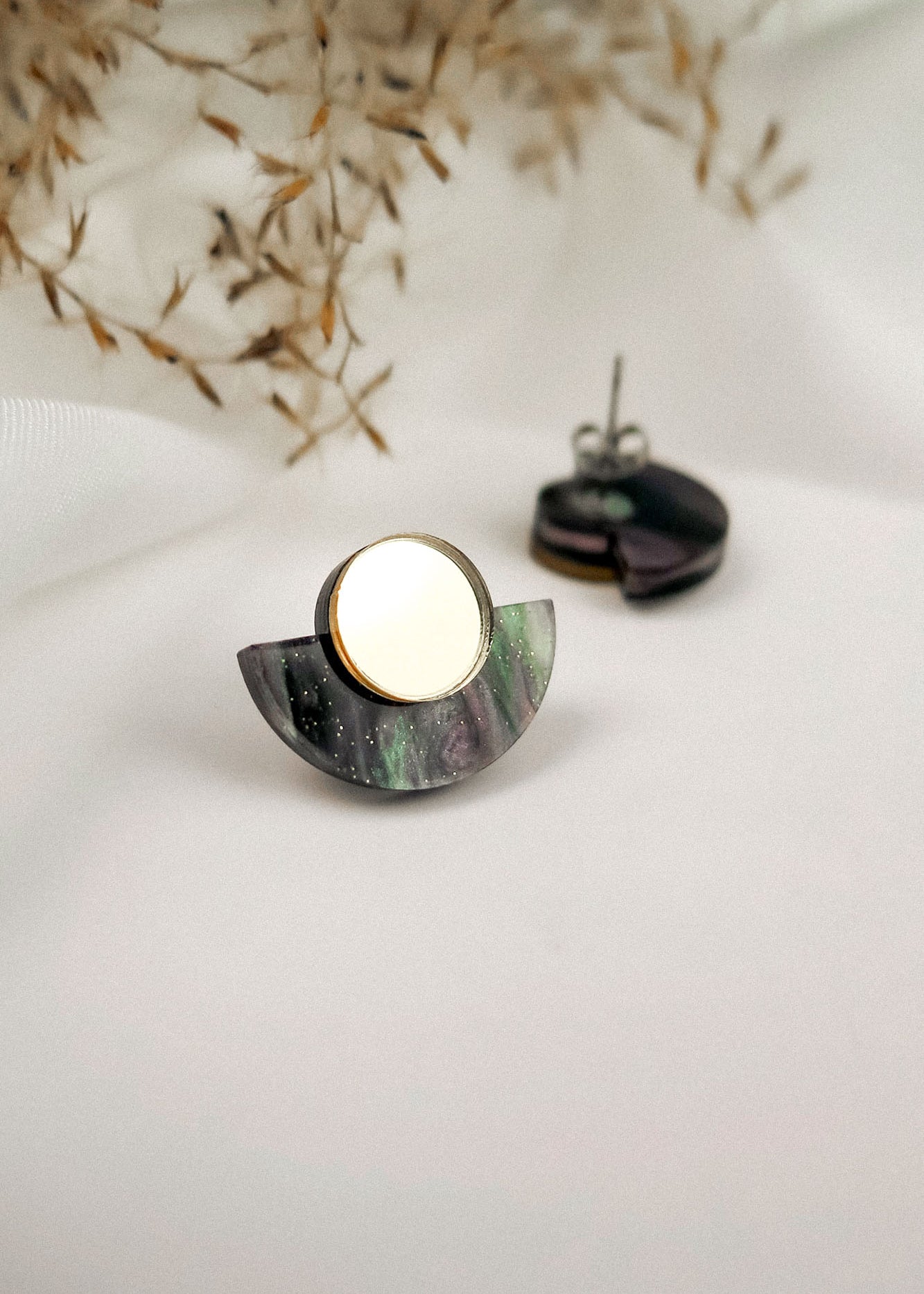 A pair of minimalist geometric stud earrings featuring a circular gold piece atop a semi-circular, marbled purple and green base, displayed on a white surface with dried foliage in the background.