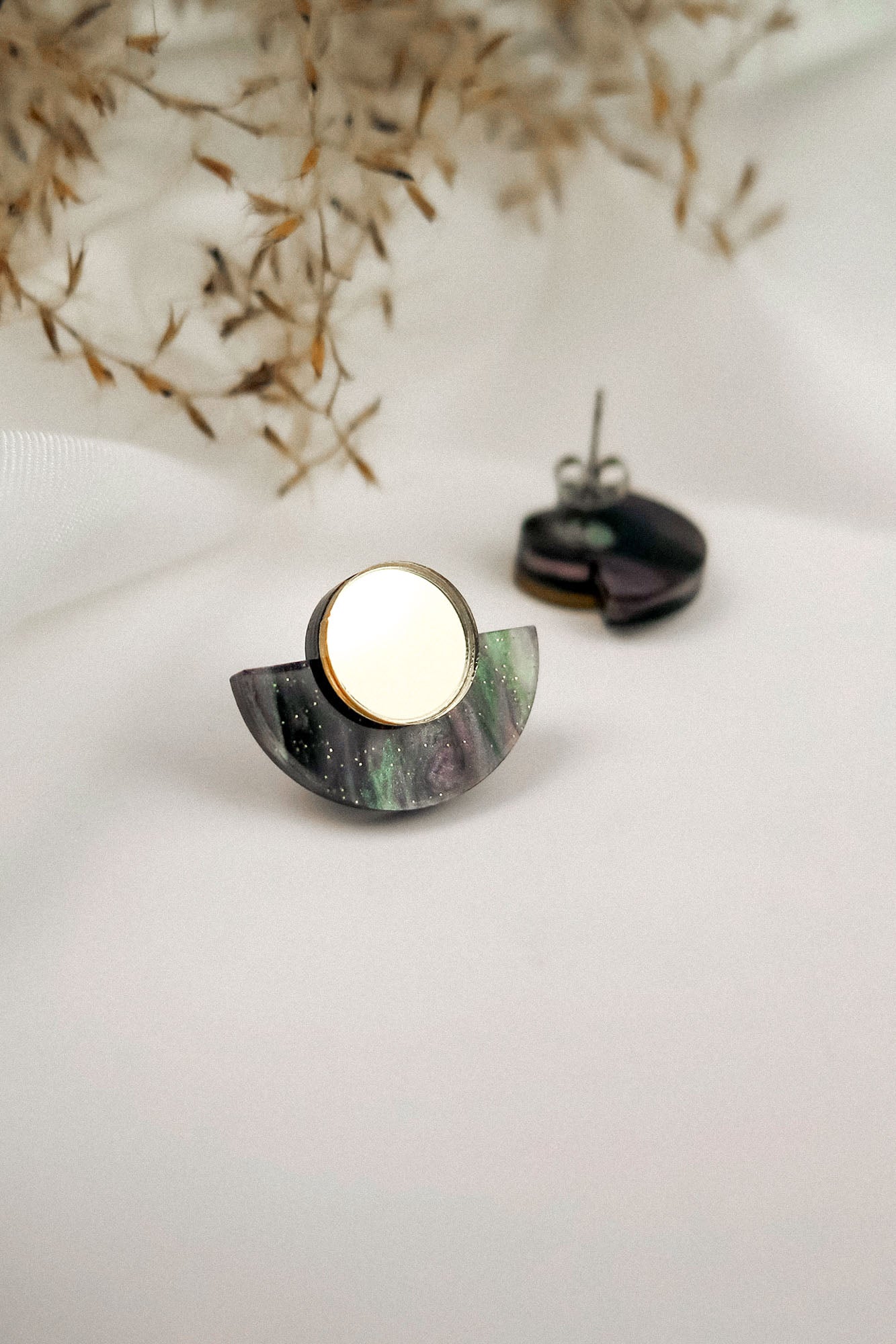 A pair of minimalist geometric stud earrings featuring a circular gold piece atop a semi-circular, marbled purple and green base, displayed on a white surface with dried foliage in the background.