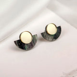A pair of round gold and black marbled minimalist geometric stud earrings with semi-circular accents, displayed on a soft white fabric background.
