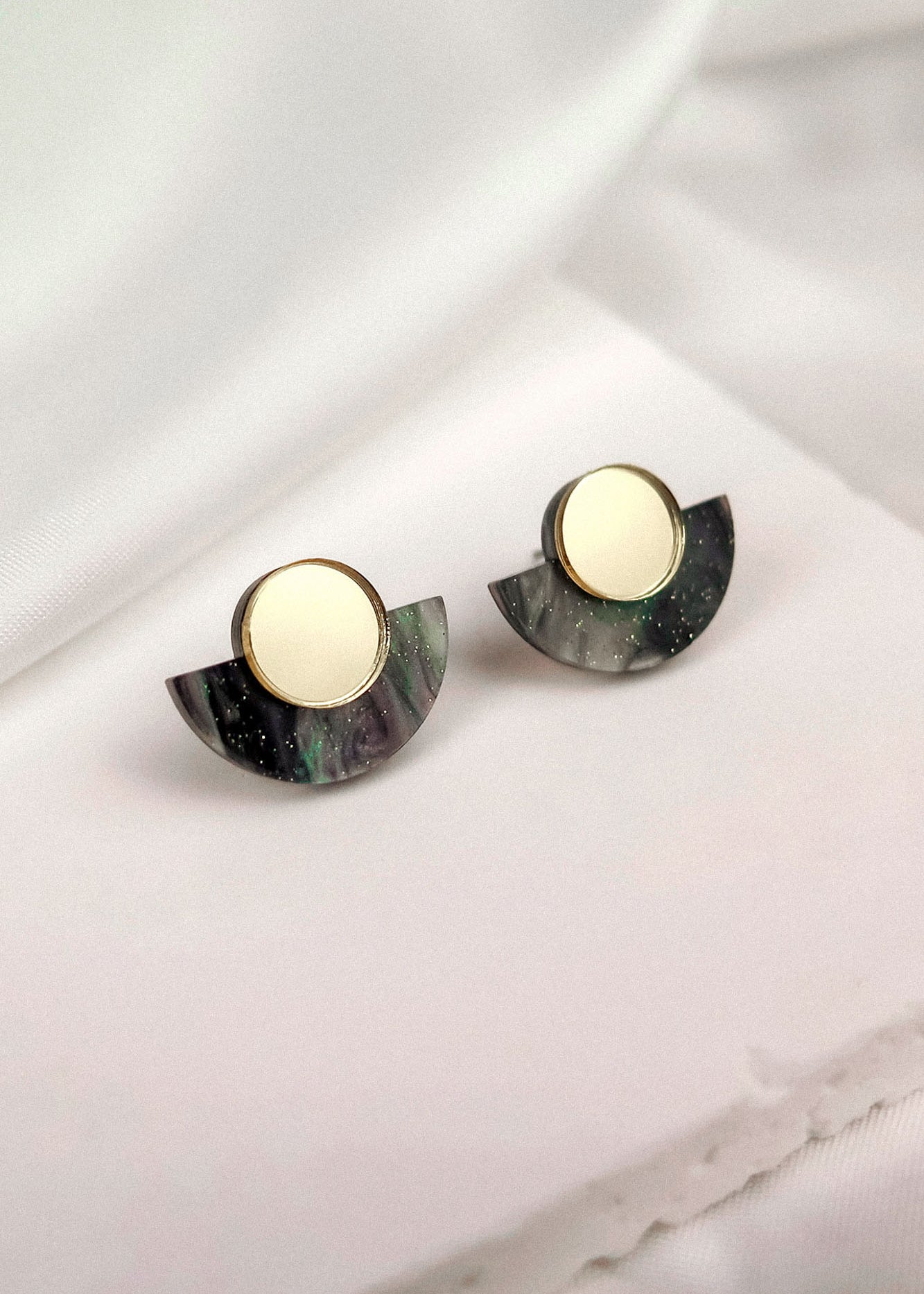 A pair of round gold and black marbled minimalist geometric stud earrings with semi-circular accents, displayed on a soft white fabric background.