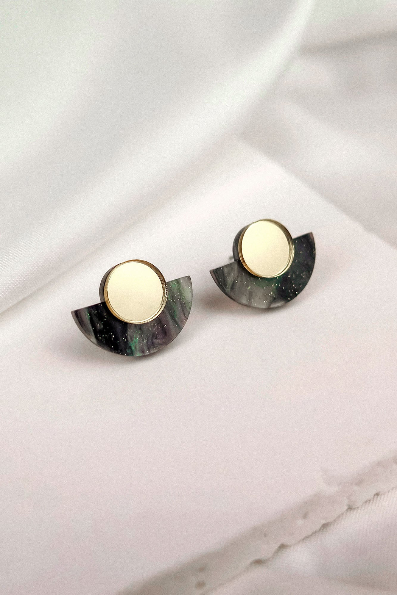 A pair of round gold and black marbled minimalist geometric stud earrings with semi-circular accents, displayed on a soft white fabric background.