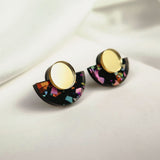 Minimalist Geometric Stud Earrings featuring round gold tops and semicircular black bases with colorful patterns. These lightweight, hypoallergenic earrings rest on a white surface with soft, light fabric in the background.