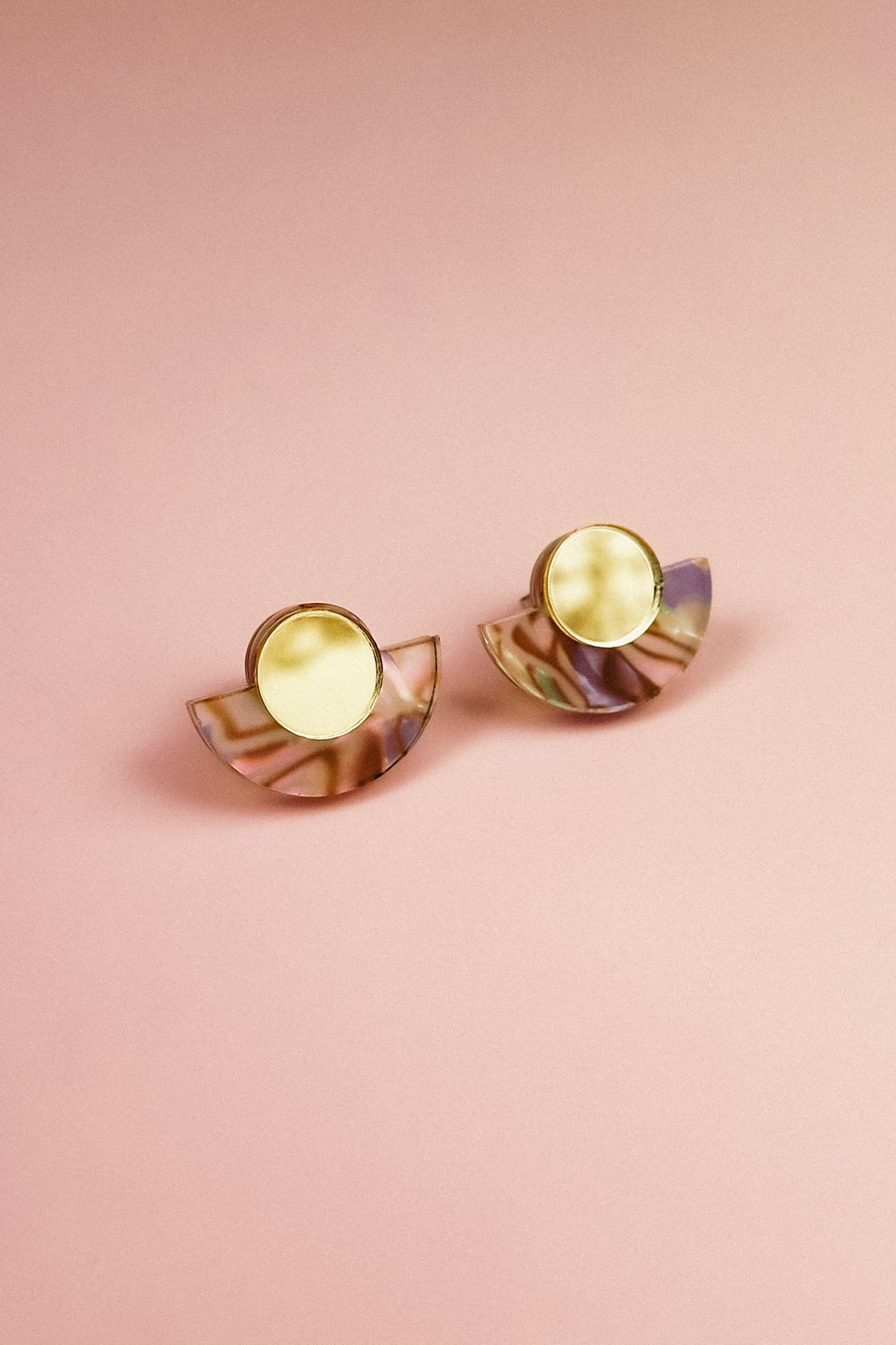 A pair of Minimalist Geometric Stud Earrings featuring round gold tops and semi-circular, iridescent bases, displayed on a pink background.