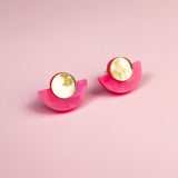 A pair of minimalist geometric stud earrings with round, gold-edged, marble-like white studs and bright pink, semicircular bases, displayed on a soft pink background. Hypoallergenic and lightweight for comfortable wear.
