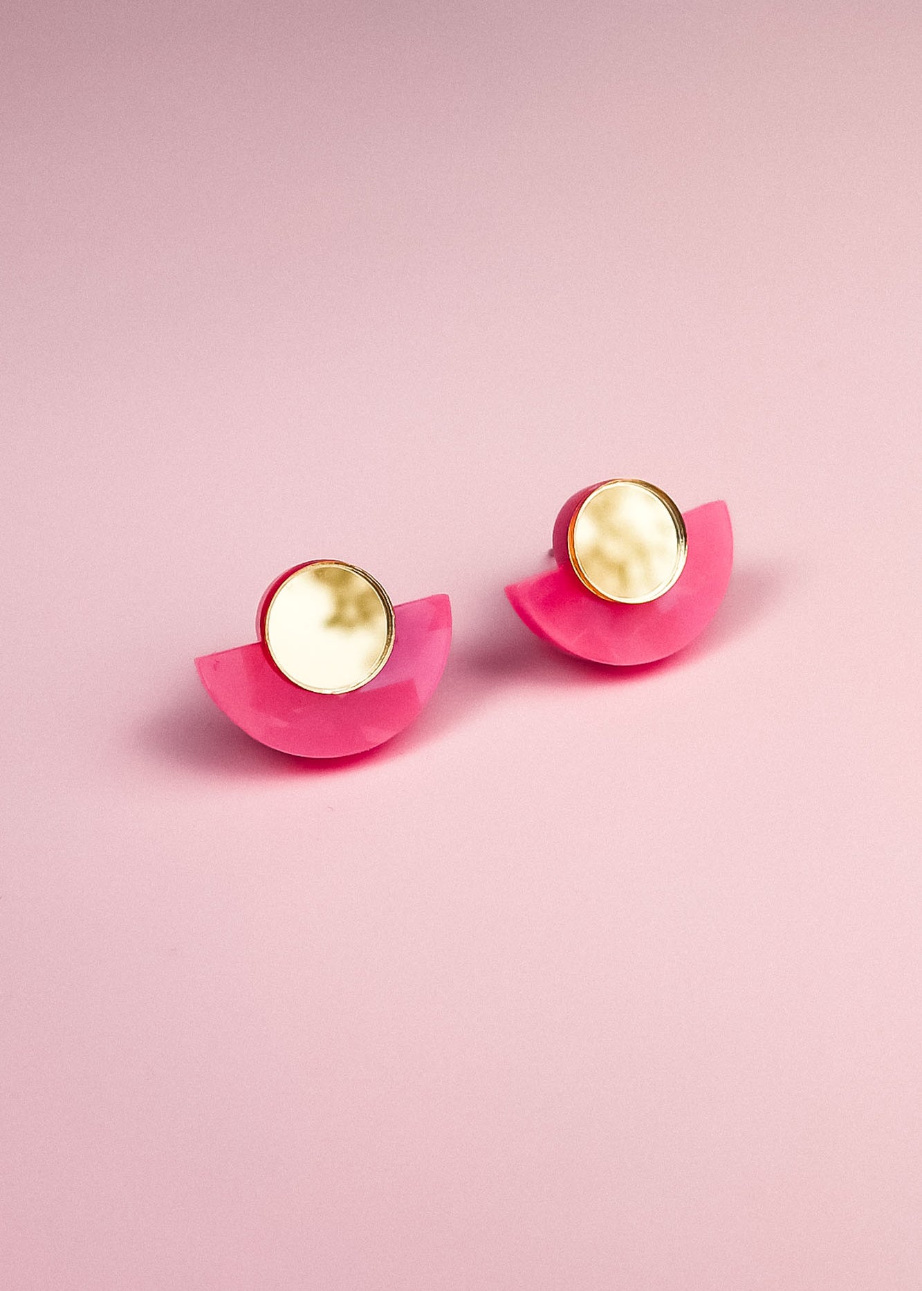 A pair of minimalist geometric stud earrings with round, gold-edged, marble-like white studs and bright pink, semicircular bases, displayed on a soft pink background. Hypoallergenic and lightweight for comfortable wear.