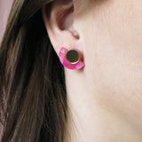 Close-up of a persons ear wearing lightweight Minimalist Geometric Stud Earrings, featuring a round gold stud and a pink, crescent-shaped accent. The person has straight brown hair tucked behind the ear.